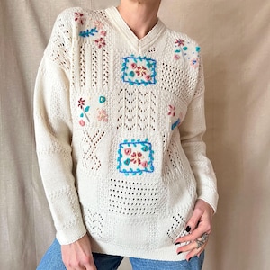 May include: Cream-colored, V-neck sweater with floral embroidery. The sweater features a mix of open knit and solid panels, with embroidered flowers in pink, blue, and green. The design includes square patches with floral motifs.