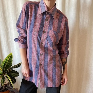 Può includere: Camicia abbottonata a righe verticali rosa e blu. La camicia ha una tasca sul petto e bottoni marroni. Le maniche sono arrotolate. Indossata con pantaloni neri.