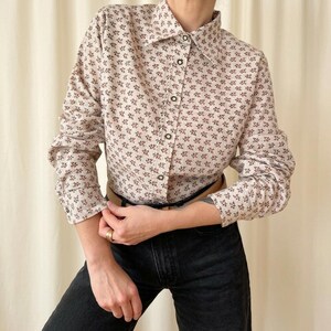 Op de afbeelding: Crèmekleurig overhemd met een bruin bloemenpatroon. Het shirt met lange mouwen heeft een klassieke kraag en wordt gedragen met een zwarte jeans en een bruine riem. Het shirt is gemaakt van een zachte, lichte stof.
