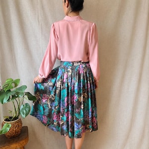 May include: A long-sleeved light pink blouse with a scalloped collar paired with a pleated midi skirt featuring a dark background and a vibrant floral pattern in shades of teal, purple, and magenta, finished with a visible back zipper closure.