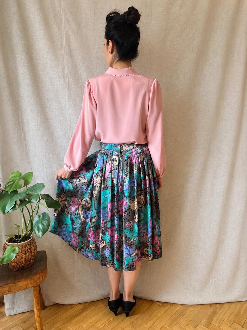 May include: A long-sleeved light pink blouse with a scalloped collar paired with a pleated midi skirt featuring a dark background and a vibrant floral pattern in shades of teal, purple, and magenta, finished with a visible back zipper closure.
