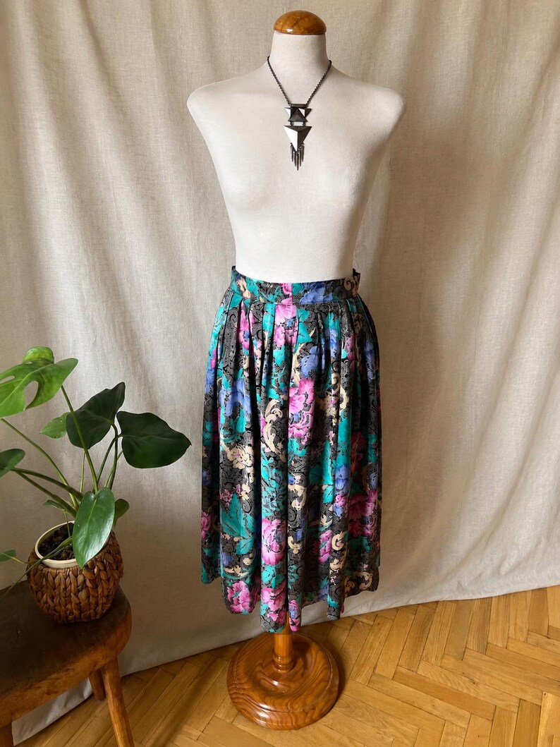 May include: A midi-length skirt featuring a vibrant floral pattern in shades of magenta, teal, and blue set against a black background with intricate gold-toned filigree details. The skirt has a high-waisted fit with soft pleats and a flared silhouette.