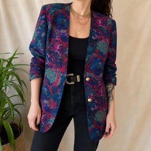 Pode incluir: Um blazer de manga comprida com um padrão abstrato vibrante em tons de magenta, azul profundo e verde-azulado. O blazer tem uma silhueta de corte reto, decote sem colarinho e dois botões circulares dourados no fecho frontal.