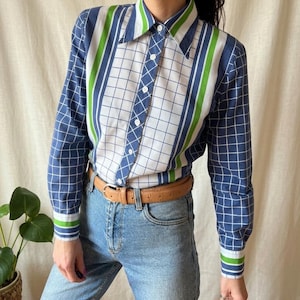 May include: A vintage button-down shirt with a striking geometric pattern. The shirt features a mix of blue grid, white, and green stripes. The long sleeves have matching striped cuffs. The shirt is paired with light blue jeans and a brown belt.