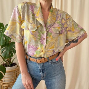 May include: A short-sleeved button-up shirt with a colorful floral and abstract pattern in shades of yellow, lavender, and green. The shirt has two front pockets and is paired with blue jeans and a brown belt.