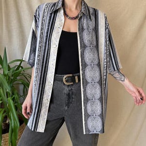May include: A short-sleeved button-down shirt featuring a vertical pattern of black, white, and grey stripes alongside a decorative panel of circular motifs. The shirt is worn over a solid black top with dark grey jeans and a black belt with a gold buckle.