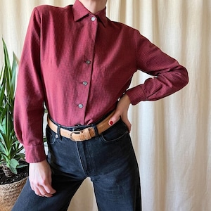 May include: A burgundy button-down shirt with a small checkered pattern. The long-sleeved shirt has a classic collar and is paired with a light brown belt and dark wash jeans. The shirt is buttoned up and has a tailored fit.