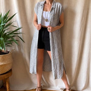 May include: A long, sleeveless, button-down duster vest featuring a grey and white snakeskin print, worn over a plain white tank top and black shorts. The duster has a collared neckline, a front button closure, and side slits at the hem.