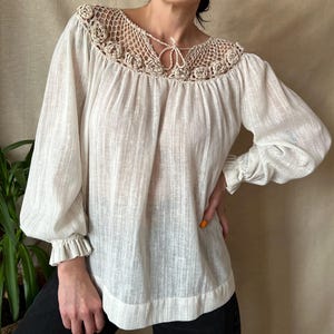 以下が含まれることがあります： オフホワイトのブラウス。かぎ針編みのネックラインと、ギャザーの入った長袖が特徴です。 かぎ針編みのディテールには花の模様があります。 ブラウスはゆったりとしたフィット感で、ギャザーの入ったネックラインです。 袖口はフリルカフスで終わっています。