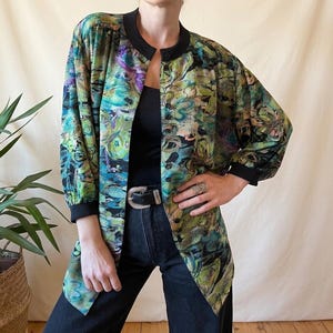 Op de afbeelding: Een jas met lange mouwen en een abstract bloemenpatroon in groen, blauw en paars. Het heeft een zwarte kraag en manchetten, gedragen over een zwarte top en donkere jeans. Een zilveren riem en ring maken de look compleet. De jas is open.