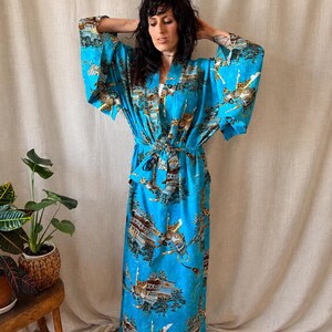 May include: A floor-length, bright blue robe with wide, loose sleeves and a matching tie belt. The fabric features a repeating print of traditional Japanese-style buildings, pine trees, and figures in armor, rendered in shades of brown, gold, and white.