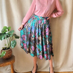 May include: A long-sleeved, light pink button-down shirt with a pointed collar and vertical pleats paired with a high-waisted, pleated midi skirt featuring a dark background with a vibrant floral pattern in shades of magenta, teal, and gold.