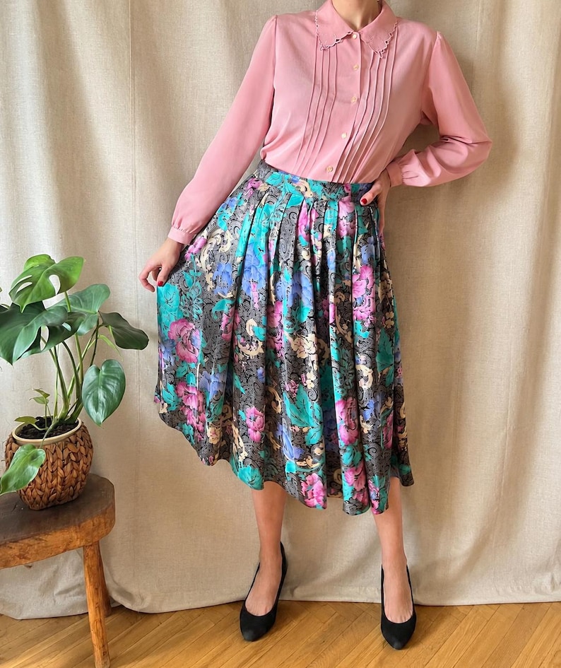 May include: A long-sleeved, light pink button-down shirt with a pointed collar and vertical pleats paired with a high-waisted, pleated midi skirt featuring a dark background with a vibrant floral pattern in shades of magenta, teal, and gold.