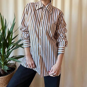 May include: A button-down shirt with vertical stripes in white, brown, and black. The long-sleeved shirt has a collared neckline and a single chest pocket. The cuffs and collar have a contrasting stripe detail. The shirt is paired with black pants.