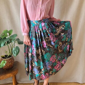 May include: A long-sleeved dusty pink button-down shirt with a scalloped collar and vertical pleats paired with a high-waisted, pleated midi skirt featuring a black background with a vibrant floral pattern in shades of pink, blue, and green.