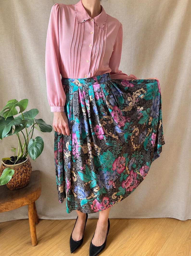 May include: A long-sleeved dusty pink button-down shirt with a scalloped collar and vertical pleats paired with a high-waisted, pleated midi skirt featuring a black background with a vibrant floral pattern in shades of pink, blue, and green.