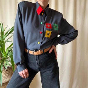 Può includere: Una camicia grigio scuro a maniche lunghe con colletto rosso e ricami quadrati colorati. La camicia presenta una cintura marrone e jeans neri. I bottoni sono rossi, verdi e gialli.