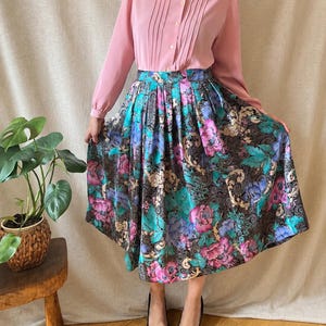 May include: A long-sleeved, light pink button-down shirt with a pointed collar and vertical pleats paired with a high-waisted, pleated midi skirt featuring a dark black background with a vibrant floral pattern in shades of blue, pink, and green.