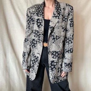Op de afbeelding: Een zwart en beige blazer met een patroon en een enkele knoopsluiting. De blazer heeft een opvallende all-over print met bloemen en geometrische ontwerpen. Het wordt gedragen met een zwarte top en een donkere jeans.