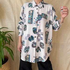 May include: White button-down shirt with a colourful abstract pattern in teal, blue, and grey. The long-sleeved shirt features a collared neckline and a button-up front. The pattern includes geometric shapes and organic forms.