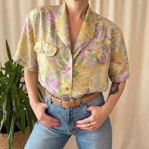 Könnte beinhalten: Kurzärmeliges Hemd mit farbenfrohem Blumenmuster in Gelb-, Lila- und Grüntönen. Das Hemd hat zwei Brusttaschen und wird mit einer blauen Jeans und einem braunen Gürtel kombiniert. Das Hemd ist aus leichtem Stoff.