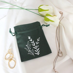 Puede incluir: Una bolsa de terciopelo verde oscuro con un diseño floral blanco. La bolsa tiene un cierre de cordón y es perfecta para guardar objetos pequeños como joyas.