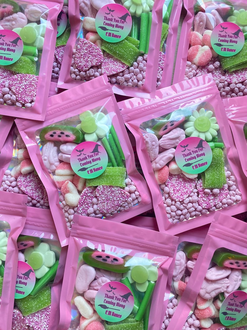 May include: Several pink cellophane bags filled with assorted candies, including pink sprinkles, green gummy sticks, and flower-shaped sweets. Each bag has a circular sticker that reads "Thank You For Coming Along" with a witch hat and broom design.