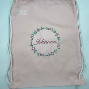May include: A pink drawstring bag with white drawstrings. The bag has a floral wreath design with the name "Johanna" embroidered in the center.
