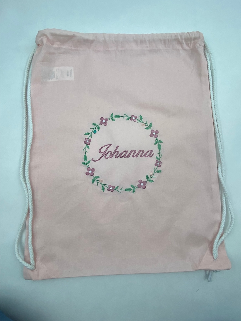 May include: A pink drawstring bag with white drawstrings. The bag has a floral wreath design with the name "Johanna" embroidered in the center.
