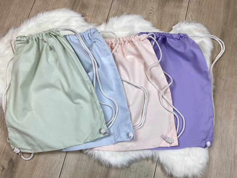 May include: Four drawstring bags in different pastel colors: light green, light blue, light pink, and lavender. The bags are made of a soft, cotton fabric and have white drawstrings.