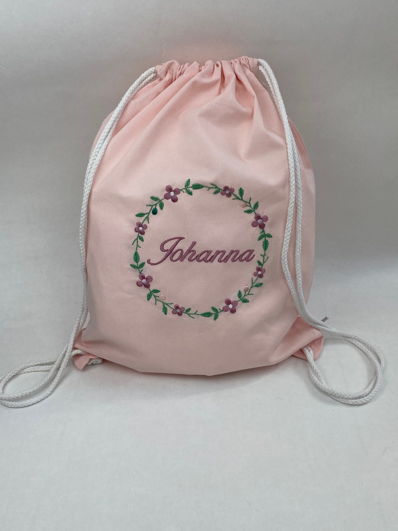 May include: A pink drawstring bag with white drawstrings. The bag has a floral wreath embroidered on the front with the name "Johanna" in the center.