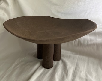 May include: A brown ceramic serving dish with a wide, shallow bowl and three short, cylindrical legs.