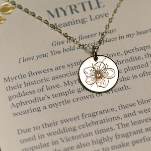 Op de afbeelding: Een gouden ketting met een ronde hanger met een delicate gegraveerde mirtebloem. De hanger wordt geleverd met een kaart die de symboliek en betekenis van de mirtebloem beschrijft: "Myrtle: Liefde. Geef de bloem om te zeggen: Ik hou van je; Je hebt een speciale plaats in mijn hart."