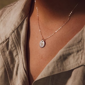 Puede incluir: Un collar de cadena de plata con un pequeño colgante redondo. El colgante tiene un diseño delicado.