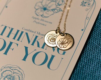May include: A gold necklace with two round pendants featuring engraved floral designs. The pendants are on a card with the text "Thinking of You" in blue lettering.