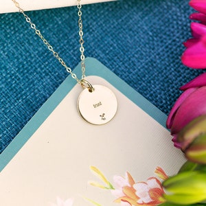 K&ouml;nnte beinhalten: Eine goldene Halskette mit einem runden Anh&auml;nger, auf dem "trust" steht. Die Kette liegt auf einem blau-wei&szlig;en Hintergrund mit rosa Blumen.
