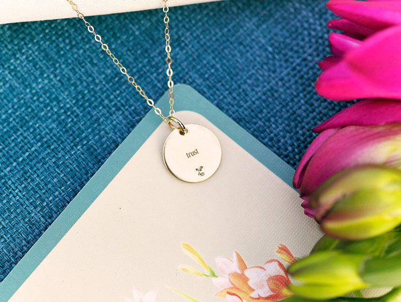 K&ouml;nnte beinhalten: Eine goldene Halskette mit einem runden Anh&auml;nger, auf dem "trust" steht. Die Kette liegt auf einem blau-wei&szlig;en Hintergrund mit rosa Blumen.
