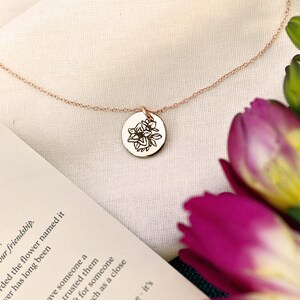 K&ouml;nnte beinhalten: Eine zarte goldene Halskette mit einem kleinen runden Anh&auml;nger, der mit einem Blumenmuster verziert ist. Der Anh&auml;nger ist mit einer Blume graviert und das Wort "Freesia" steht auf einer Karte neben der Halskette. Die Karte enth&auml;lt auch die Bedeutung der Blume, "Vertrauen".