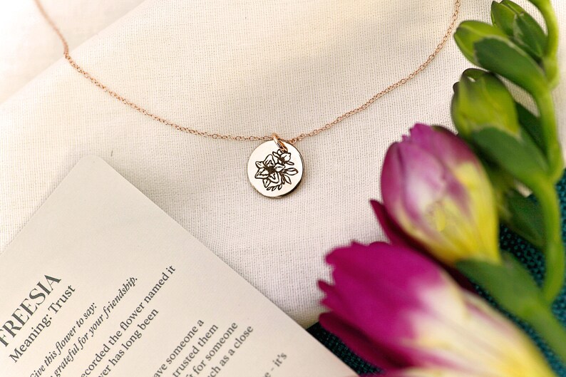 K&ouml;nnte beinhalten: Eine zarte goldene Halskette mit einem kleinen runden Anh&auml;nger, der mit einem Blumenmuster verziert ist. Der Anh&auml;nger ist mit einer Blume graviert und das Wort "Freesia" steht auf einer Karte neben der Halskette. Die Karte enth&auml;lt auch die Bedeutung der Blume, "Vertrauen".