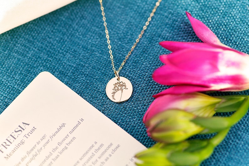 K&ouml;nnte beinhalten: Eine goldene Halskette mit einem runden Anh&auml;nger, der eine zarte, gravierte Freesie zeigt. Der Anh&auml;nger befindet sich auf einem blauen Hintergrund mit rosa Freesien.