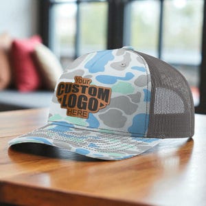 May include: A trucker hat with a camouflage pattern in shades of blue, gray, and white. The hat features a brown leather patch with the text "Your Custom Logo Here". The back of the hat is a dark gray mesh.