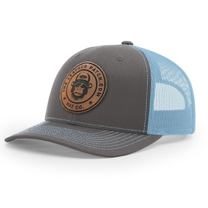 May include: A charcoal gray baseball cap with a light blue mesh back and light blue stitching on the brim. A circular brown leather patch is centered on the front, featuring a graphic of a gorilla wearing a hat and the text THE LEATHER PATCH.COM HAT CO.