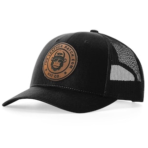 May include: A black baseball cap with a mesh back and a circular tan leather patch on the front. The patch features a graphic of a monkey wearing a hat and the text THE LEATHER PATCH.COM HAT CO. with two small stars on the sides of the circular border.
