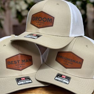 May include: Three beige and white trucker hats with white mesh backs. Each hat features a brown leather-like hexagonal patch. The patches are engraved with the text GROOM, BEST MAN, or GROOMSMAN, along with the date 9.27.2025 and a small icon.