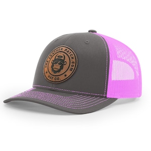 May include: A charcoal gray trucker hat with a bright neon pink mesh back and matching pink stitching on the brim. A circular brown leather patch is centered on the front, featuring a graphic of a monkey wearing a hat and the text THE LEATHER PATCH.COM HAT CO.