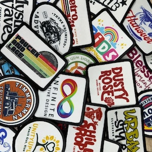 May include: A collection of embroidered patches with various designs and text. Patches include a rainbow, a shrimp, the words "Dusty Rose," and "Urban Style." The patches are in various shapes and sizes, with black borders and a mix of colors.
