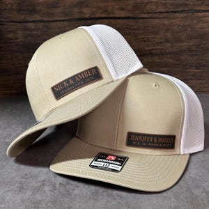May include: Two beige trucker hats with white mesh backs. Each hat features a rectangular brown leather patch. One patch reads NICK & AMBER October 17th, 2026. The other patch reads JENNIFER & MARCUS XI.X.MMXXVI and has a Richardson 112 sticker on the brim.