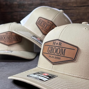 May include: Three beige trucker hats with white mesh backs. Each hat features a brown leather-like patch with black text. The front patch displays M+R, GROOM, and the date 04.25.2026. Other patches show BEST MAN and GROOMSMAN with the same date.