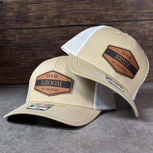 May include: Two beige and white trucker hats with mesh backs. Each hat features a brown leather-like patch with a black horizontal stripe. One patch reads D+R GROOM 07.08.2026, and the other patch reads D+R BRIDE 07.08.2026 in black lettering.