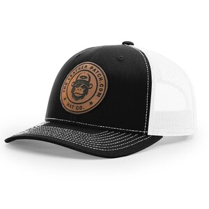 May include: A black and white trucker hat featuring a circular tan leather patch on the front. The patch displays a graphic of a monkey wearing a hat and sunglasses, surrounded by the text THE LEATHER PATCH.COM HAT CO. with white stitching.
